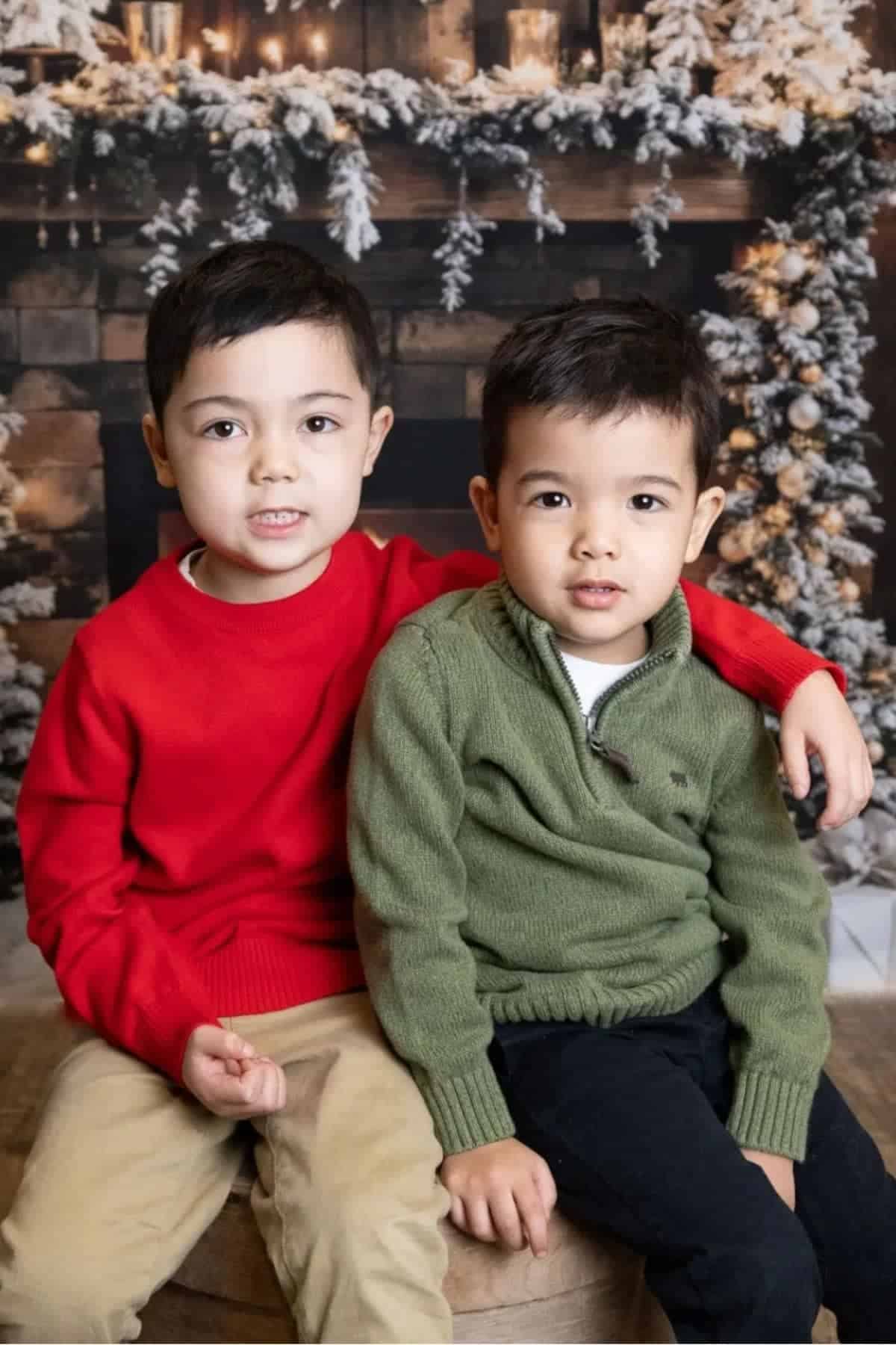 Two young boys sit closely together in front of a festive, winter-themed backdrop with garlands and lights. One wears a red sweater; the other wears a green sweater.