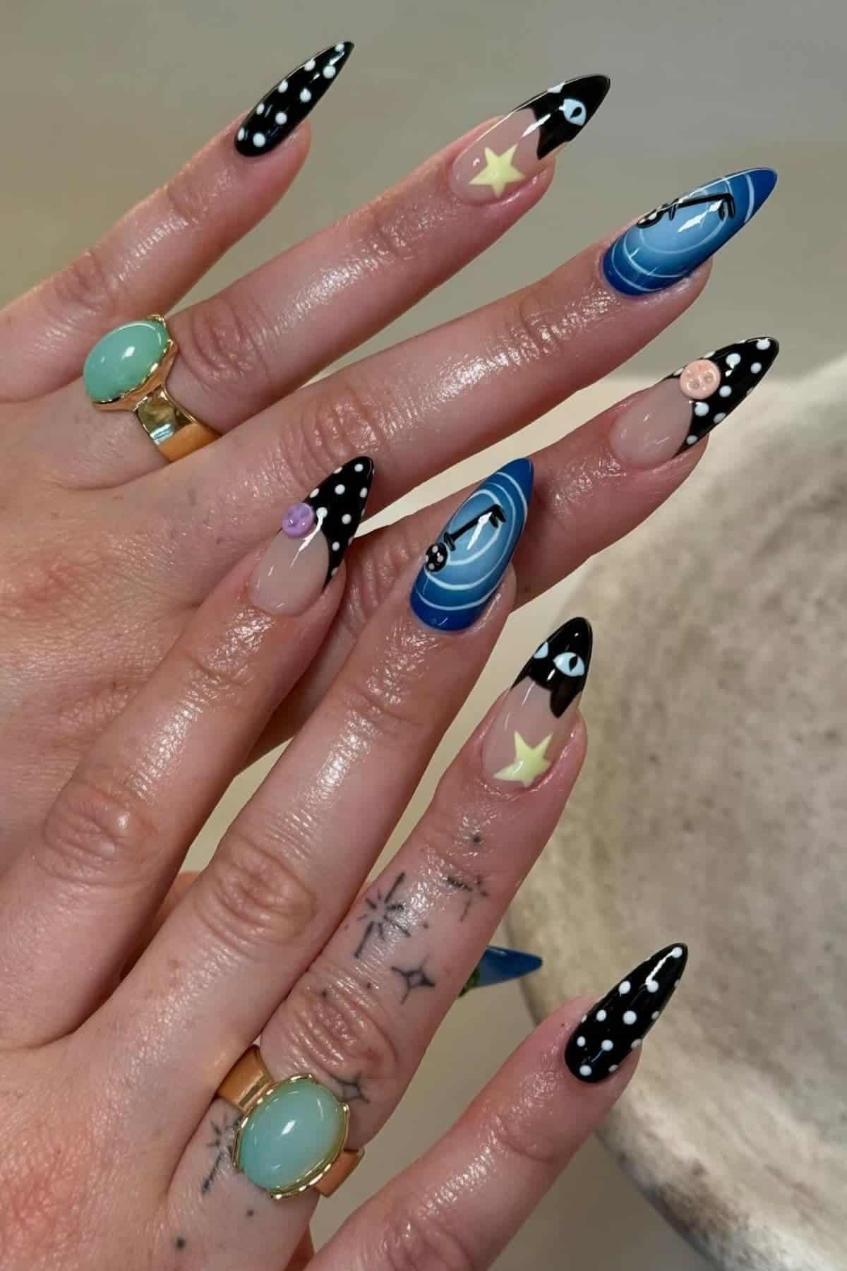 Hands with long, pointed nails featuring black, white, and blue celestial designs, including stars, eyes, planets, and dots. The person is wearing green rings and has small tattoos on their fingers.