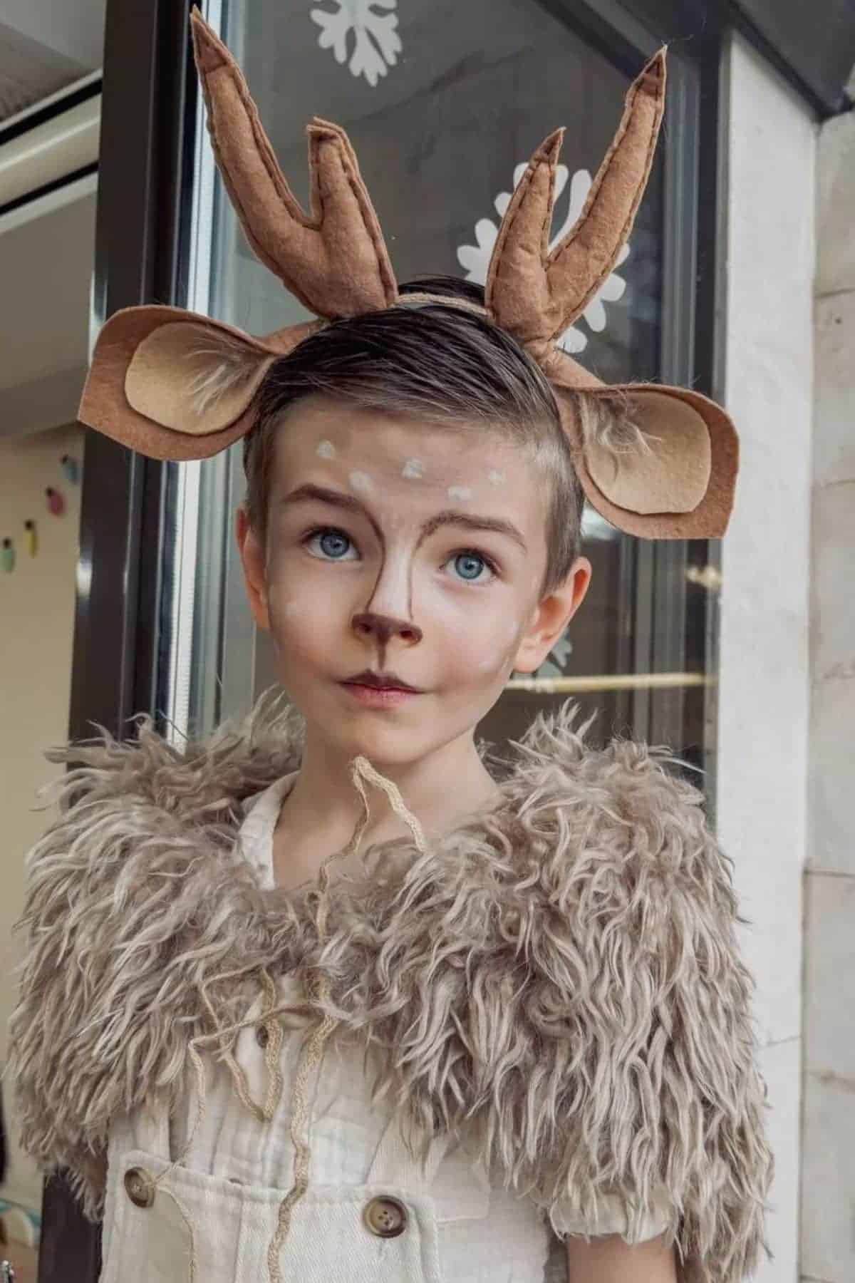 A child dressed as a deer with antler headband, ear accessories, brown fur shawl, and face painted with deer-like features, standing indoors near a window.