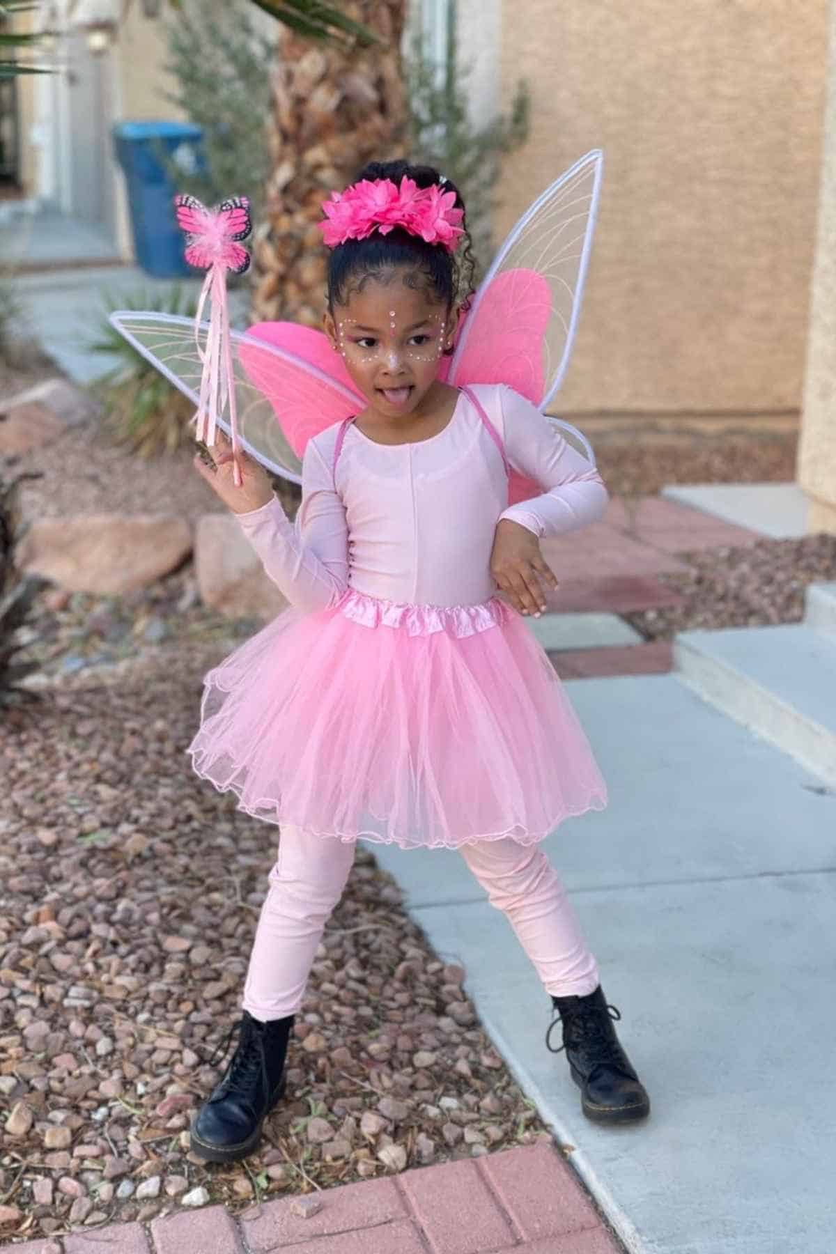 Young child in a pink fairy costume with wings and wand poses outdoors on a sidewalk next to rocks and plants.