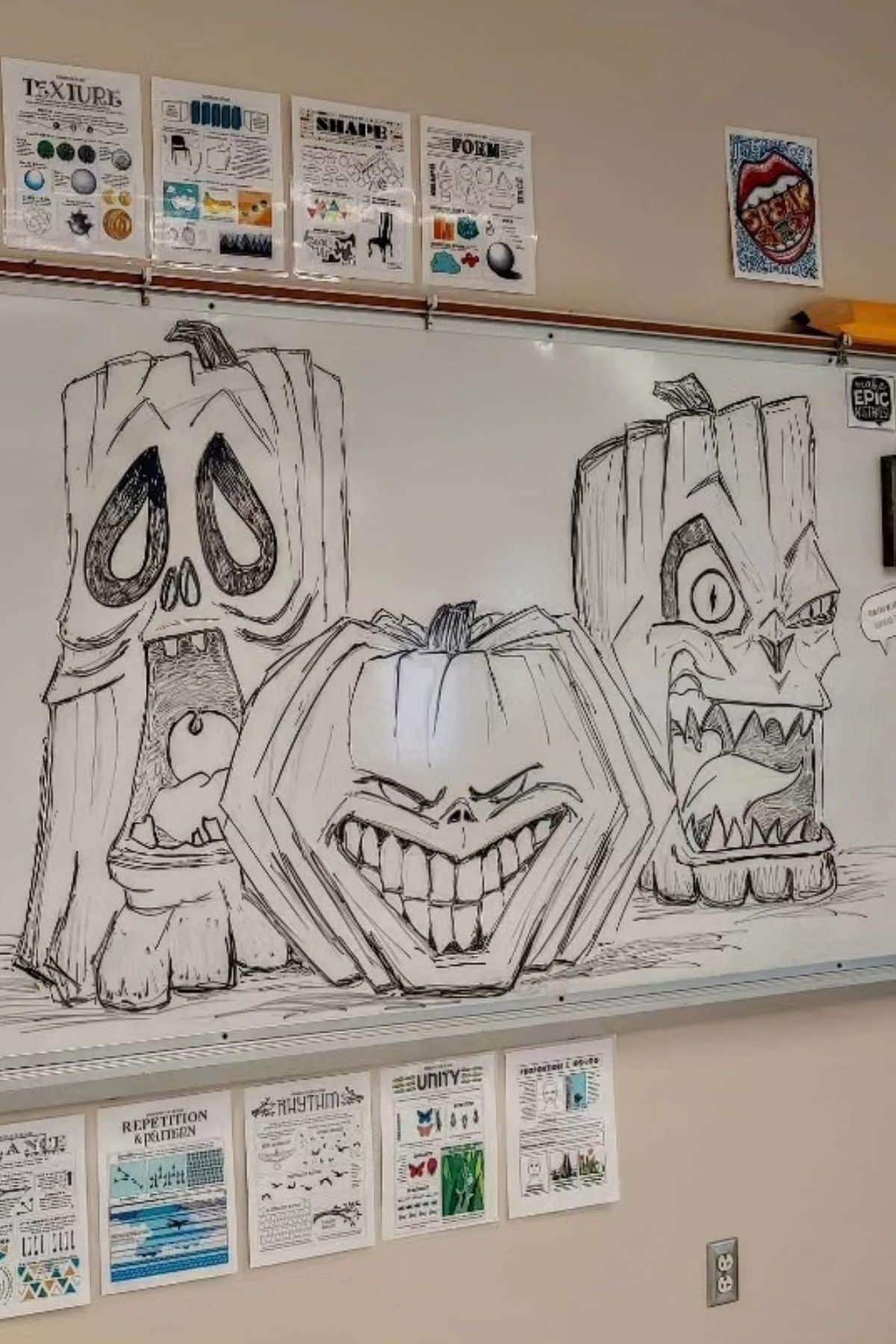 Three cartoonish pumpkins with exaggerated expressions are drawn in black marker on a classroom whiteboard, surrounded by educational posters about art elements and principles.