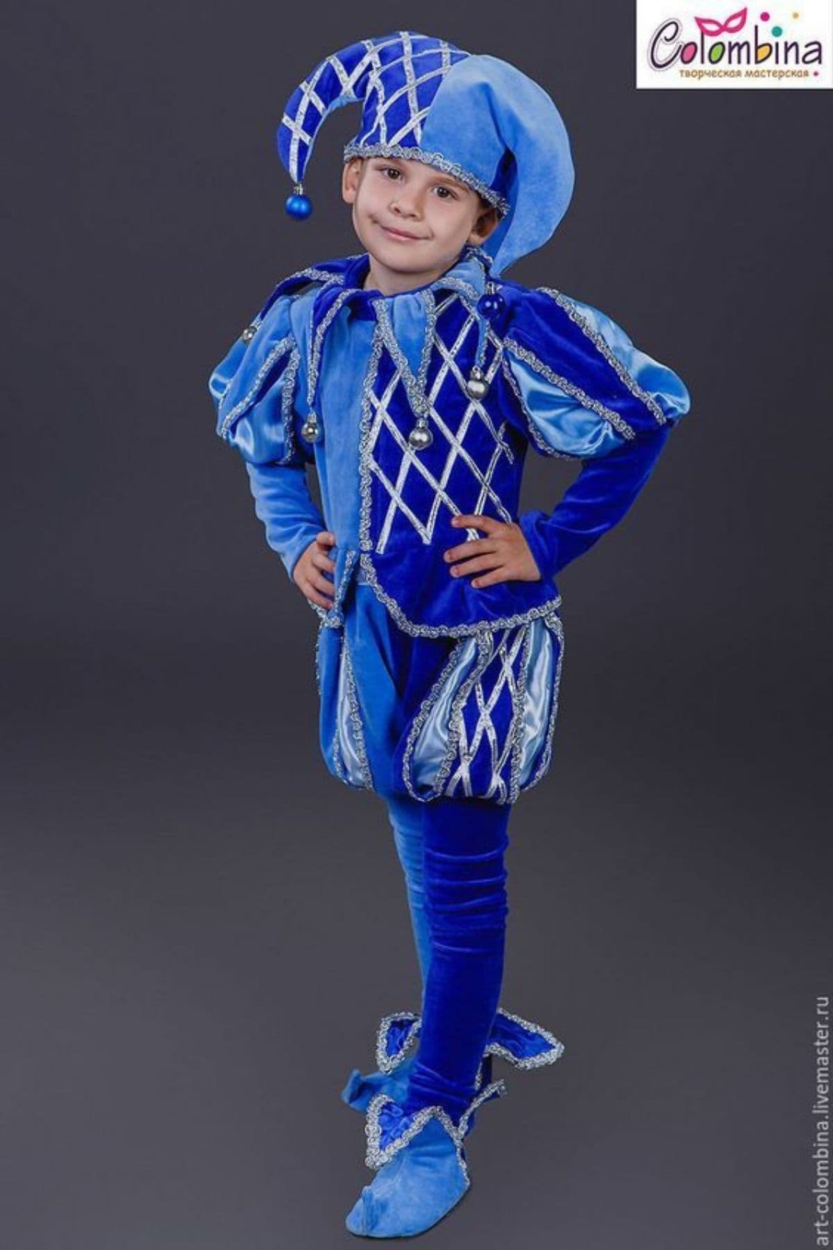 A child wearing a bright blue jester costume with a matching hat, standing on a dark background with hands on hips.
