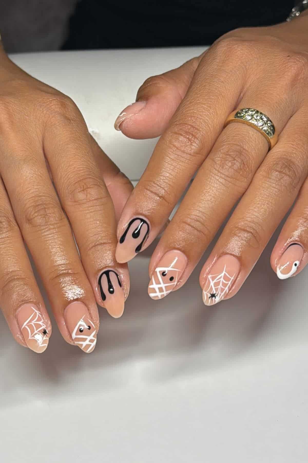 Hands with nude-colored nail polish featuring black drip, white spiderweb, and eye designs; gold ring on left hand.