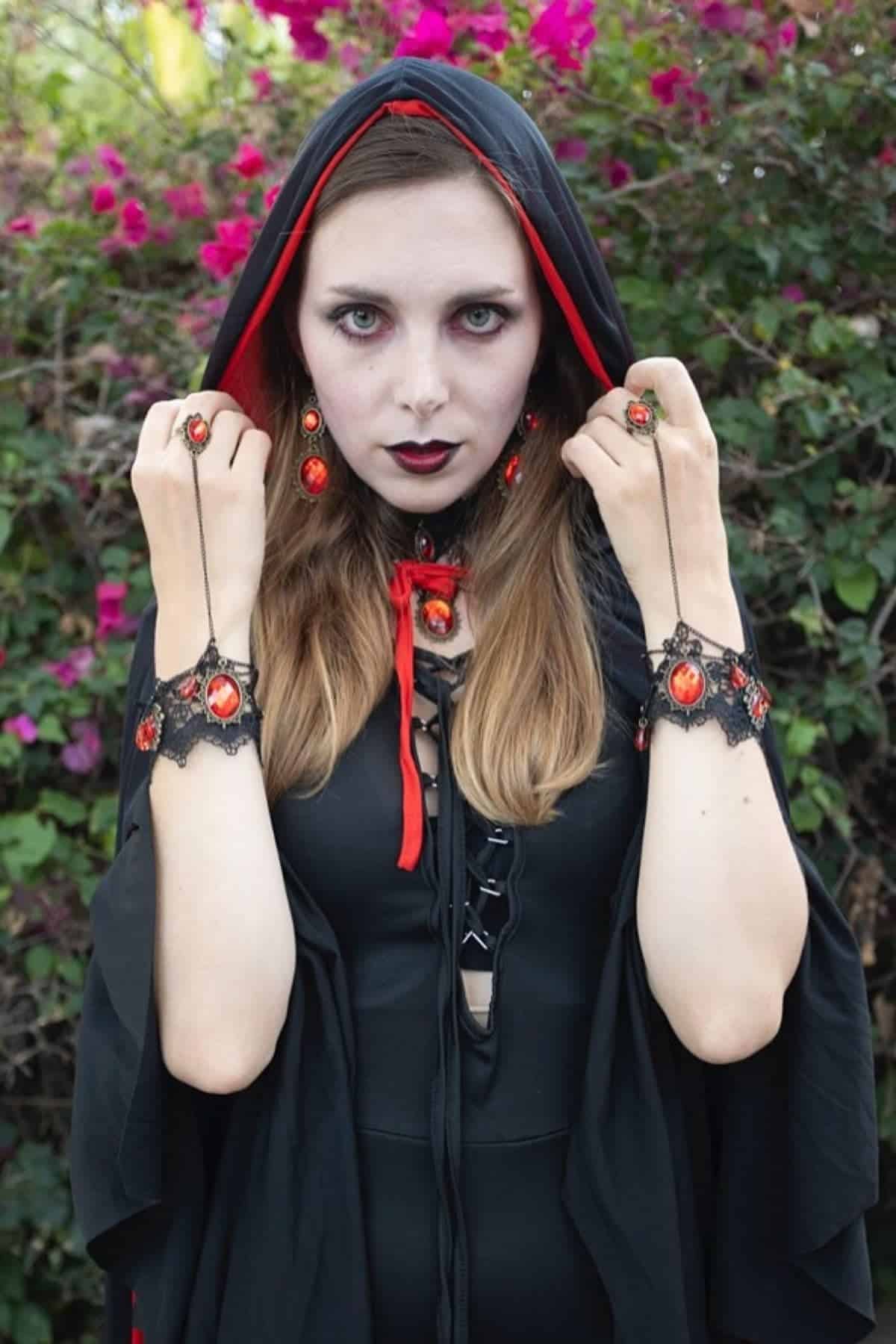 A woman in a black and red hooded costume with gothic jewelry stands outdoors in front of flowering bushes, looking directly at the camera.