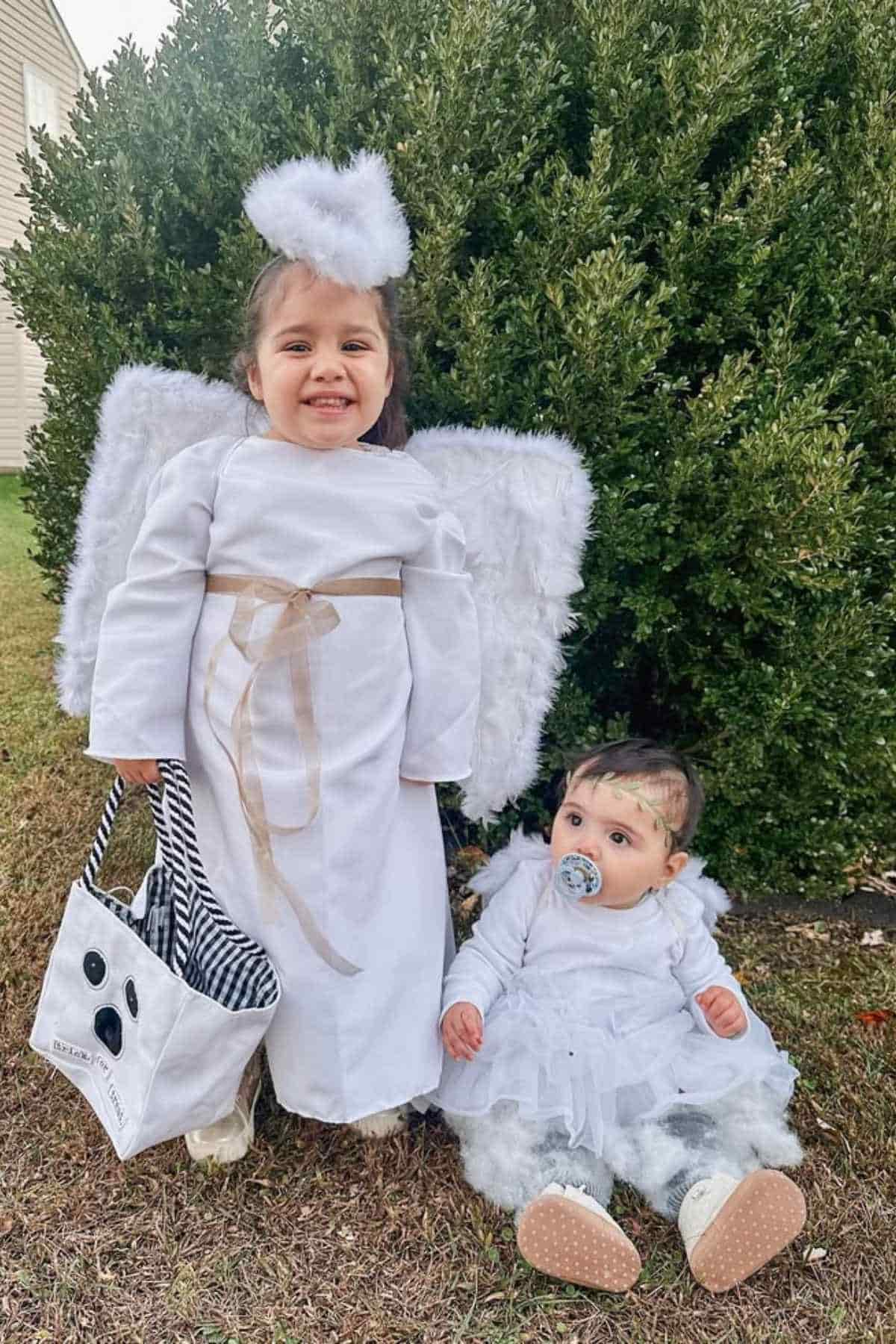 Two young children dressed as angels pose outdoors; one stands holding a ghost-themed treat bag, while the other sits on the grass with a pacifier, both wearing white costumes and wings.