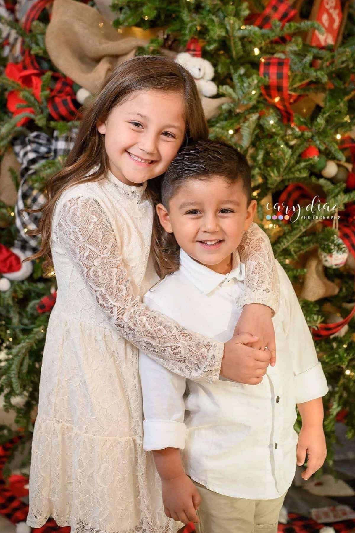 Two children dressed in white pose in front of a decorated Christmas tree, smiling for the camera. The girl stands behind the boy with her arms around his shoulders.