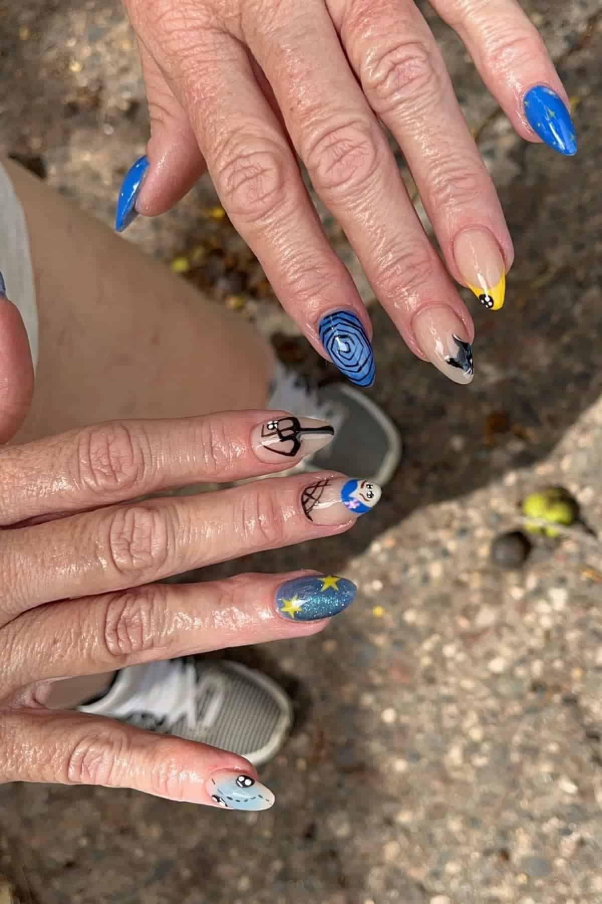 Hands with long, pointed nails featuring blue polish, swirl patterns, spider webs, stars, and abstract designs. The background is an outdoor, rocky ground with part of a shoe visible.