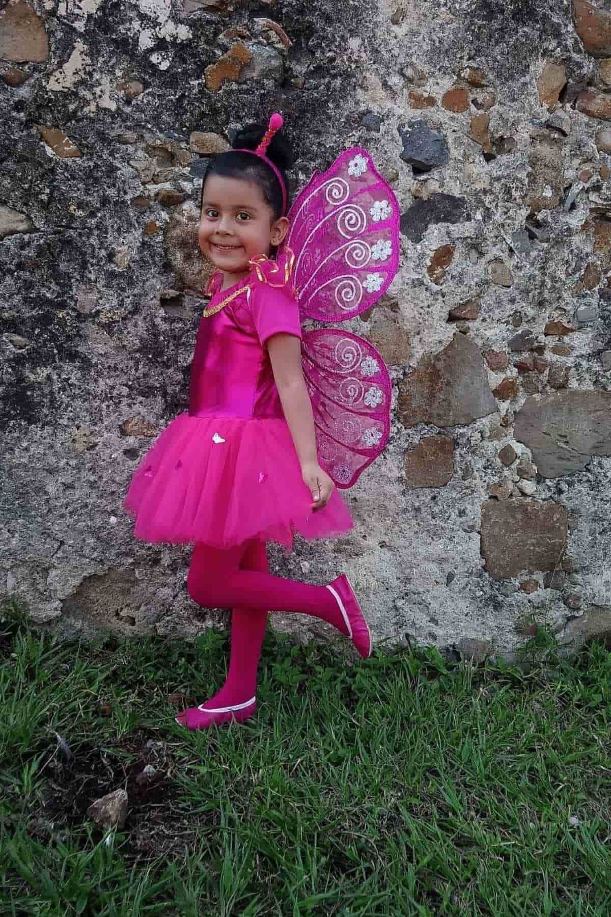 Young child in a bright pink fairy costume with wings stands on grass near a stone wall, smiling and posing with one foot raised.