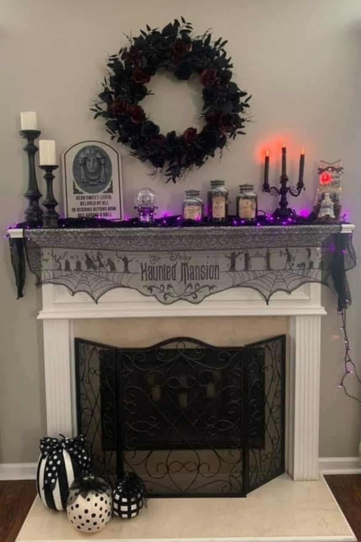 A fireplace decorated for Halloween with black candles, jars, a wreath, purple lights, and a “Haunted Mansion” sign. Black and white pumpkins are placed on the floor in front.