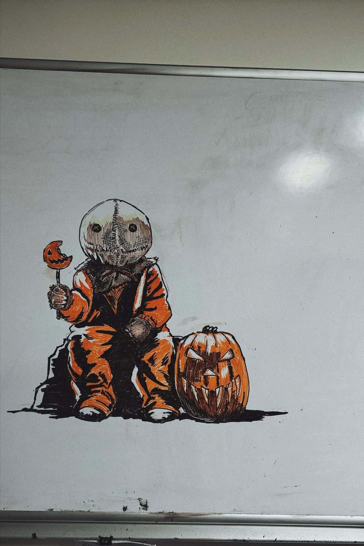 Drawing of a child in an orange costume holding a pumpkin lollipop, sitting next to a carved jack-o’-lantern, on a whiteboard.