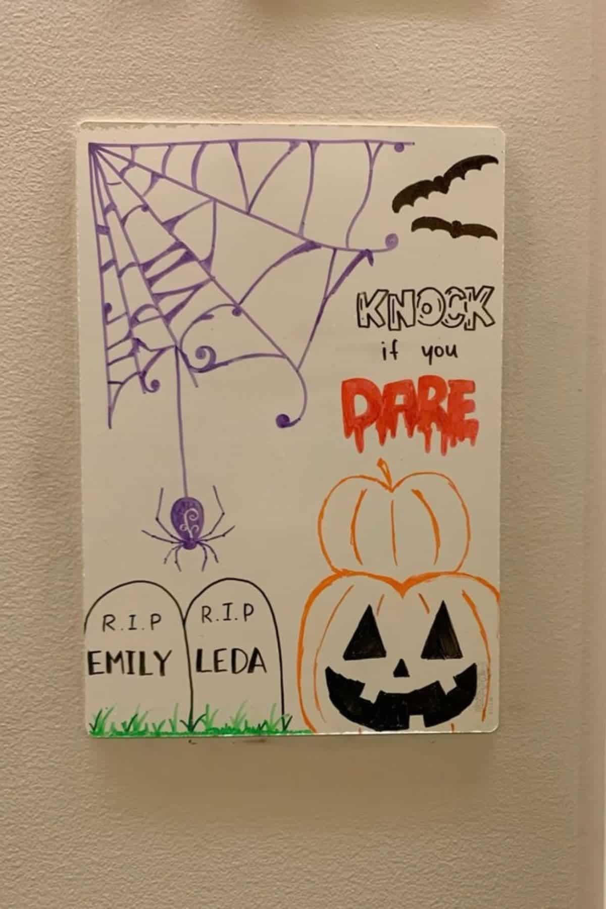 A whiteboard displays Halloween drawings: a spider web, spider, bats, two gravestones labeled “Emily” and “Leda,” the words “Knock if you dare,” and a jack-o’-lantern.