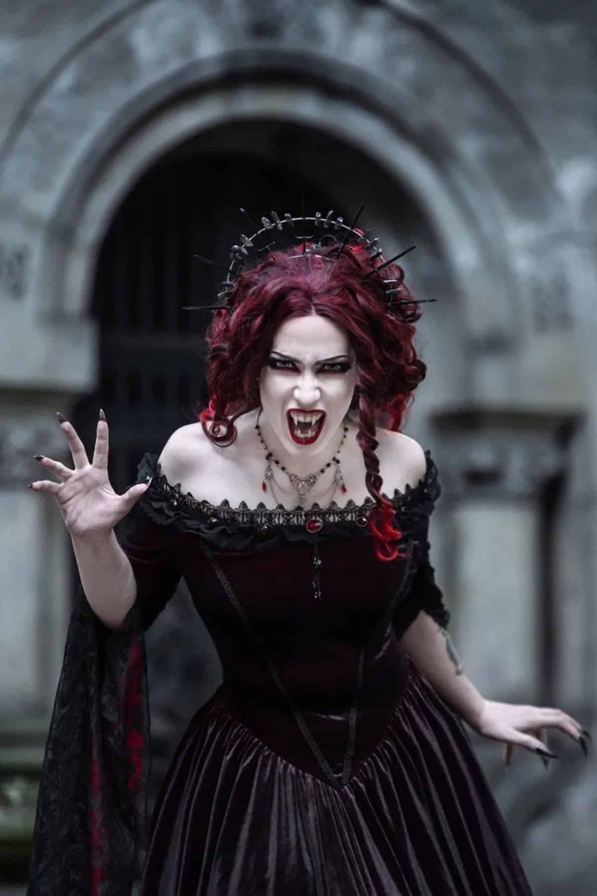 Person in gothic vampire costume with fangs and claw-like hand gesture, wearing a dark dress and ornate headpiece, posing in front of a stone archway.