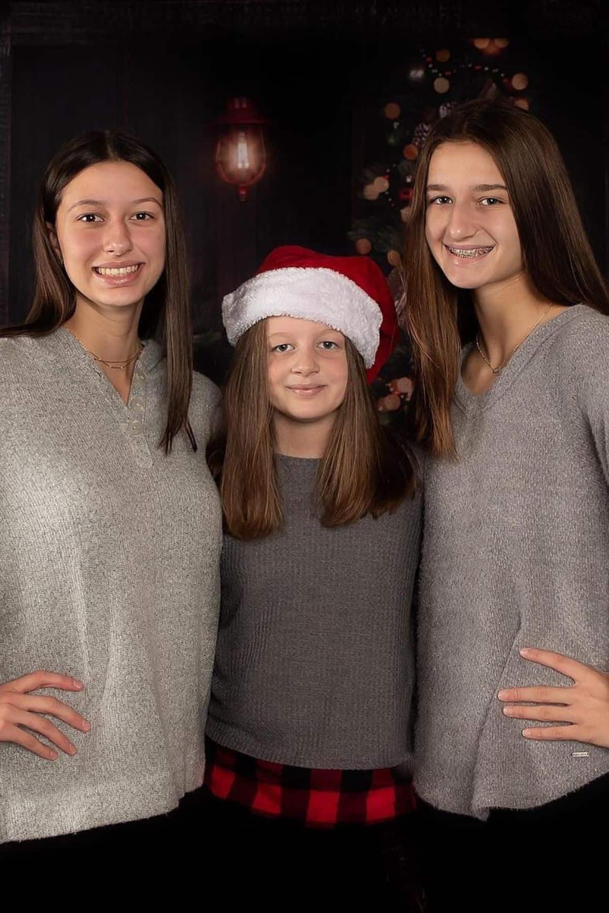 Three girls stand together indoors; the middle girl wears a Santa hat, all wear gray sweaters, and a decorated Christmas tree is visible in the background.