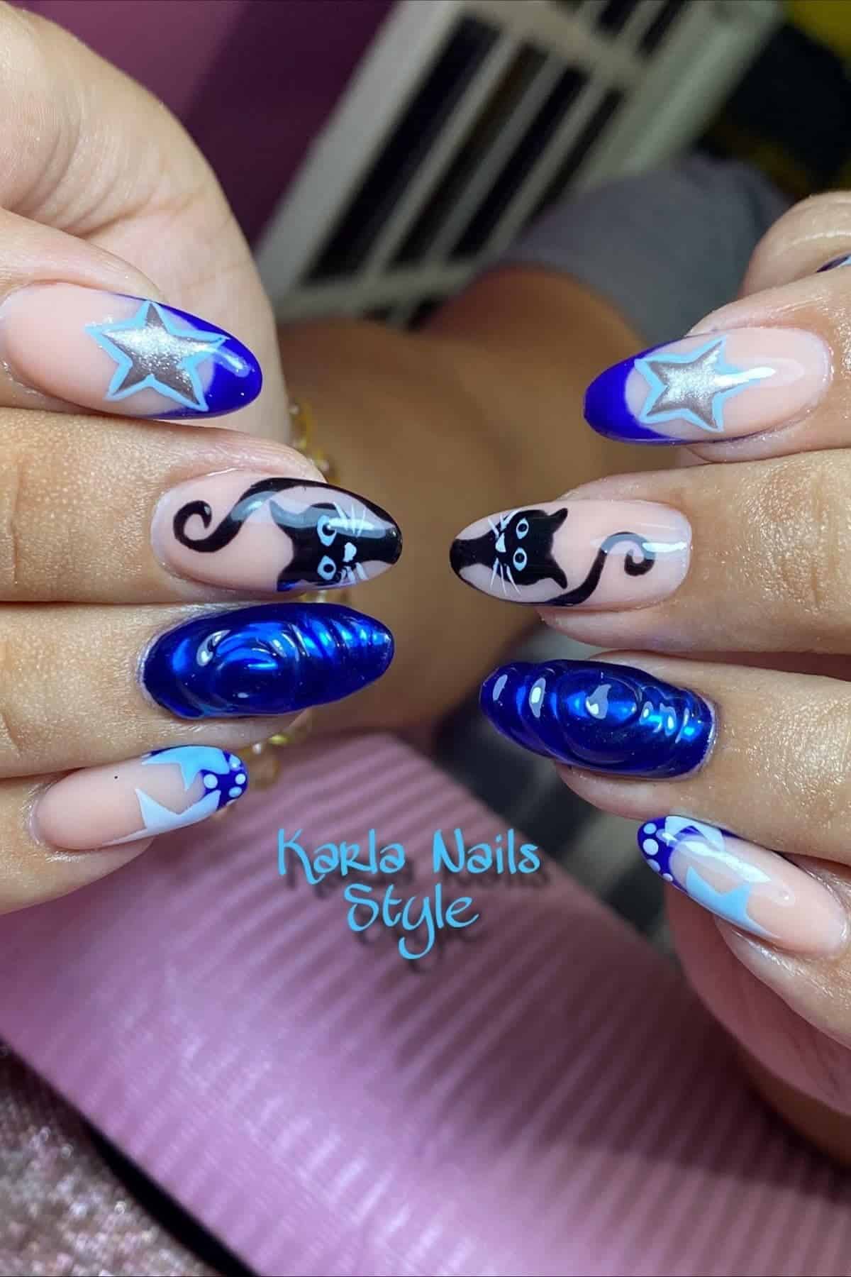 Hands with long almond-shaped nails featuring blue stars, 3D blue swirls, and black cat designs on a nude base. Text “Karla Nails Style” is visible below the nails.