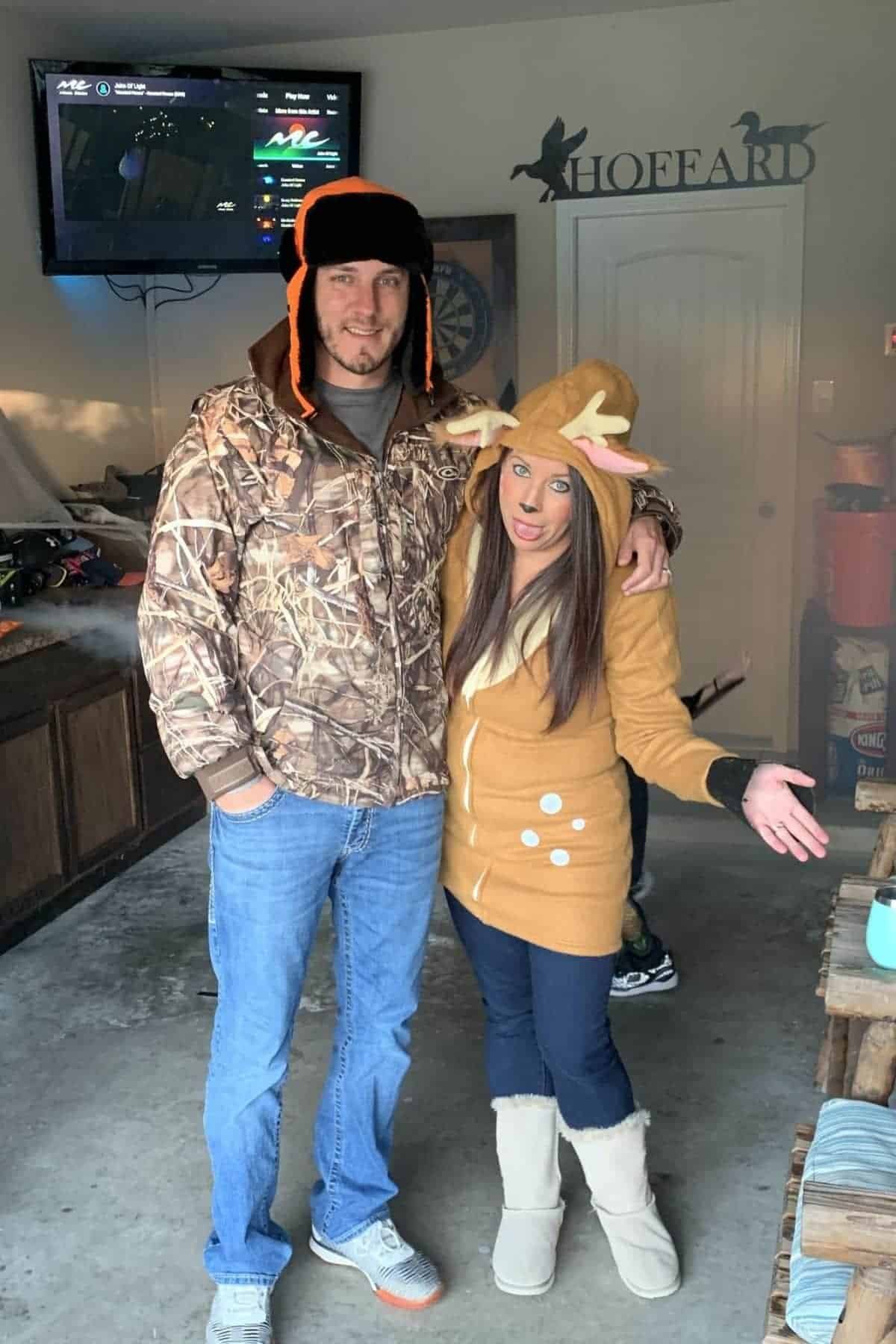 A man in a camo jacket and a woman in a reindeer hoodie pose together in a garage. A TV is mounted on the wall behind them.