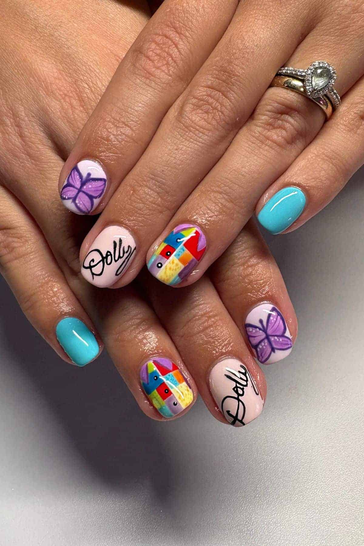 Hands with light blue and pastel pink nails, featuring purple butterflies, “Dolly” text, and a colorful abstract face design. The person is wearing rings, including a diamond ring.