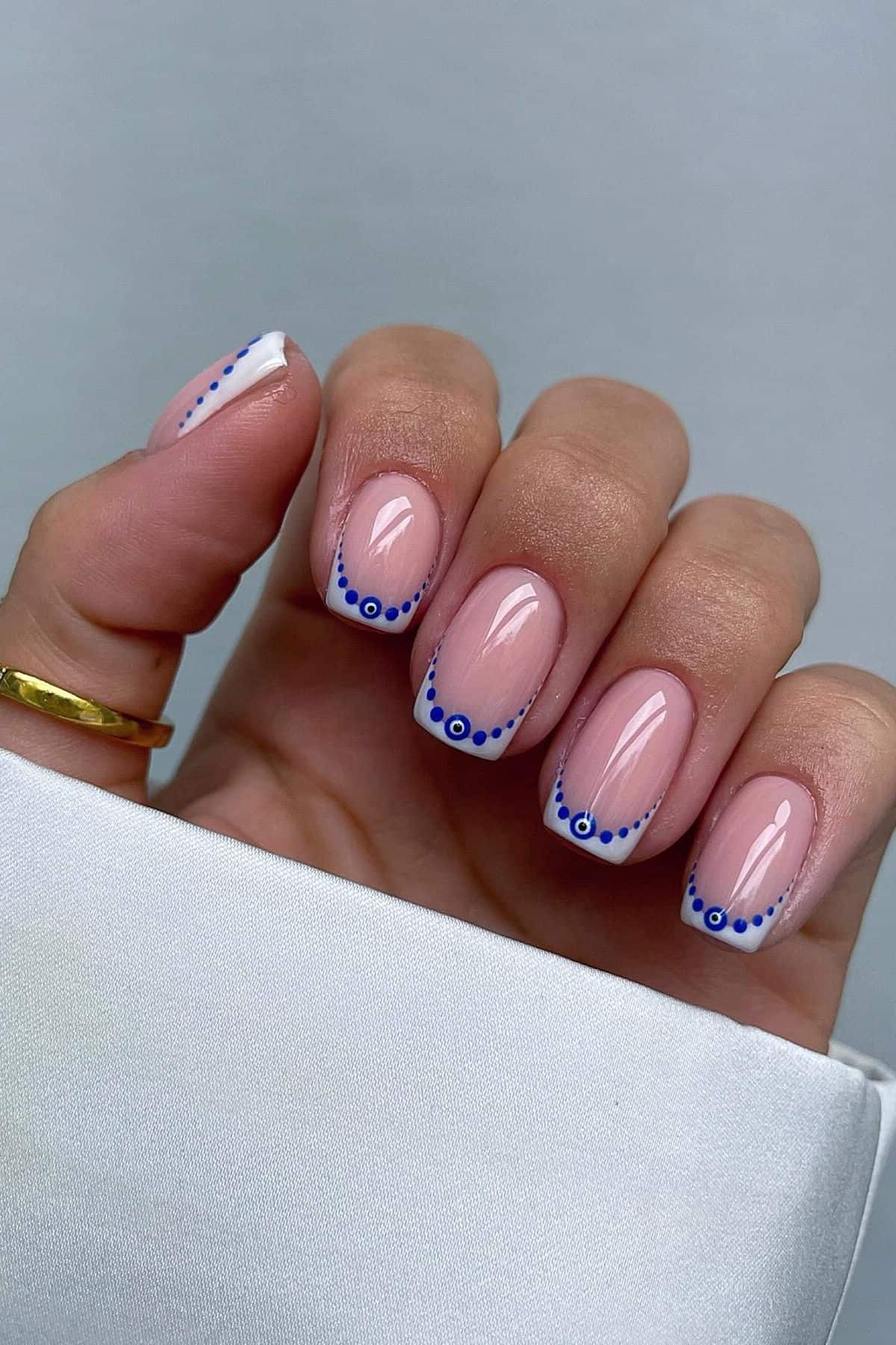 A hand with a French manicure featuring white tips and small blue dot designs on each nail, shown against a neutral background.