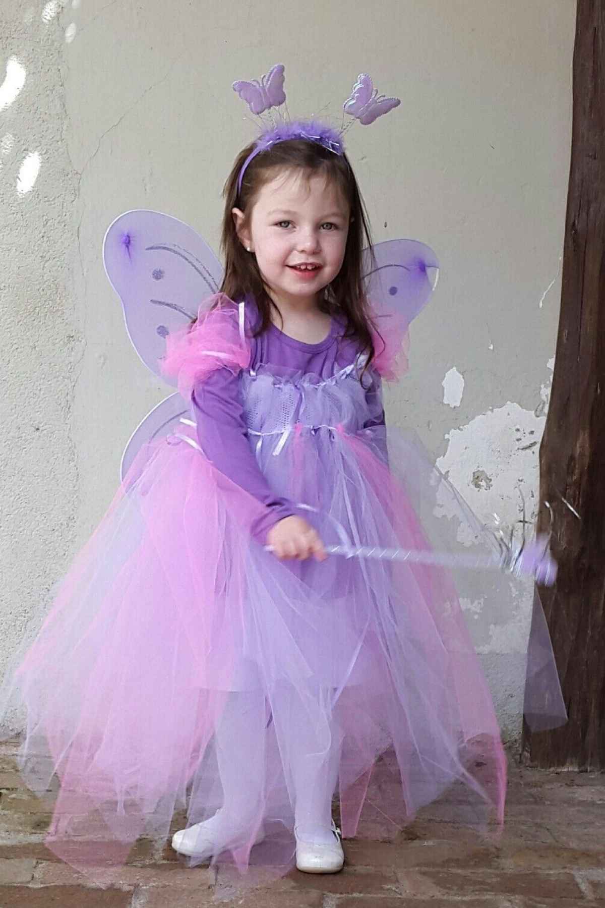 Young child stands smiling in a purple fairy costume with wings, a tutu, a headband with antennae, and holding a matching wand, against a lightly textured wall and wooden post.