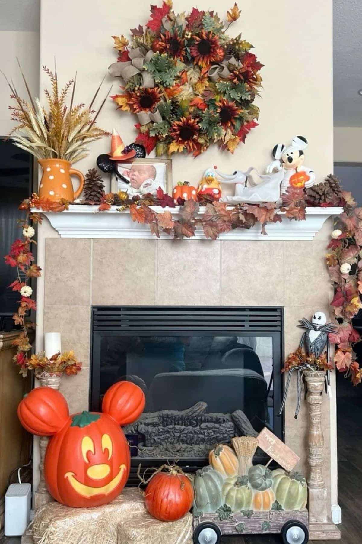 Fireplace decorated for autumn with a wreath, pumpkins, Mickey Mouse pumpkin, fall leaves, stuffed animals, scarecrow, and harvest-themed items on the mantel and hearth.