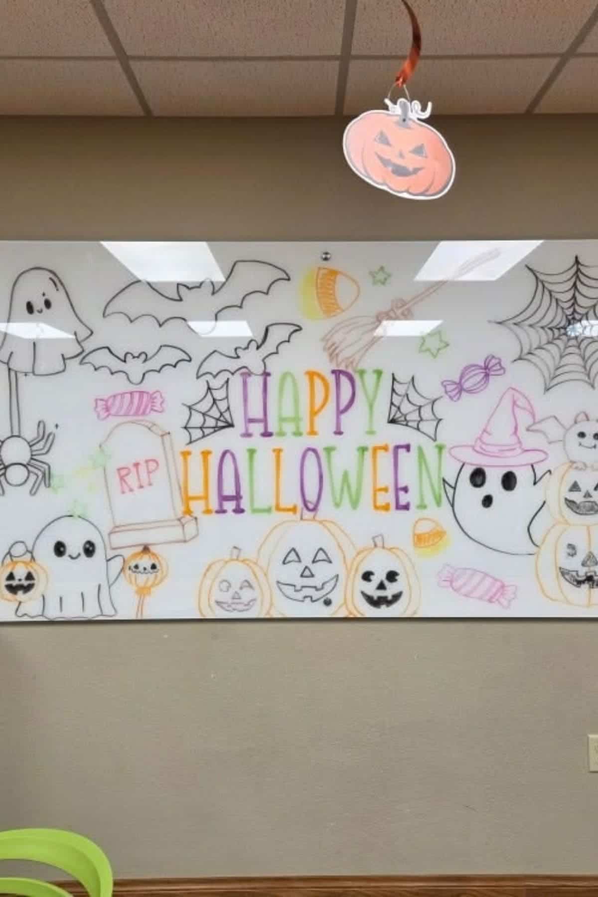 Whiteboard with “Happy Halloween” in colorful letters, surrounded by hand-drawn ghosts, bats, pumpkins, spider webs, and candy. A paper pumpkin decoration hangs from the ceiling.