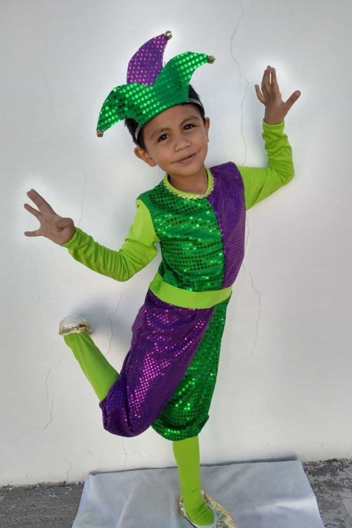 Child wearing a sparkly green and purple jester costume poses on one foot against a plain white wall, arms raised and smiling.
