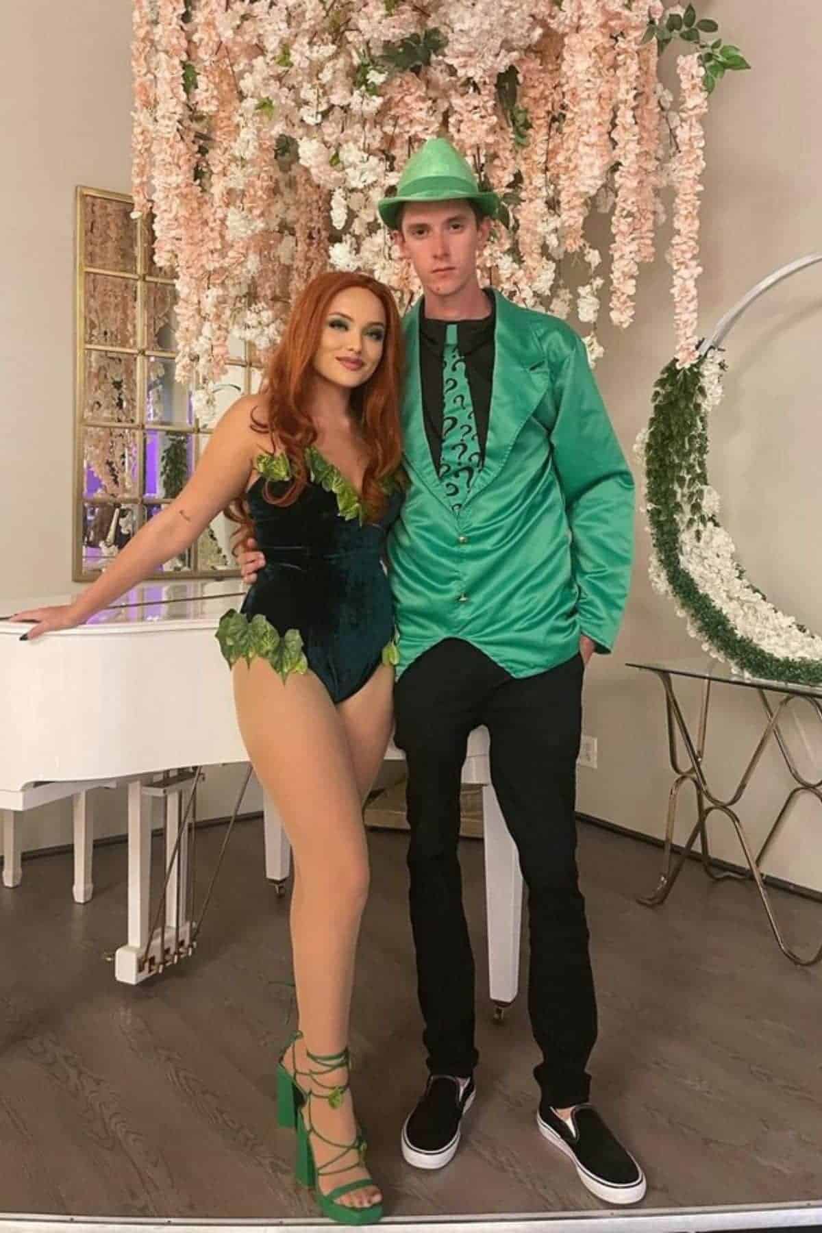 Two people in costume pose indoors; one wears a green leafy bodysuit and heels, the other wears a green suit jacket, hat, and black pants. Pink flowers hang above them.