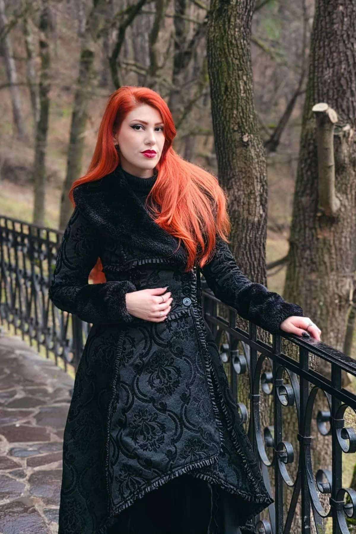 Woman with long red hair in a black patterned coat stands by a wrought iron fence in a forested area.