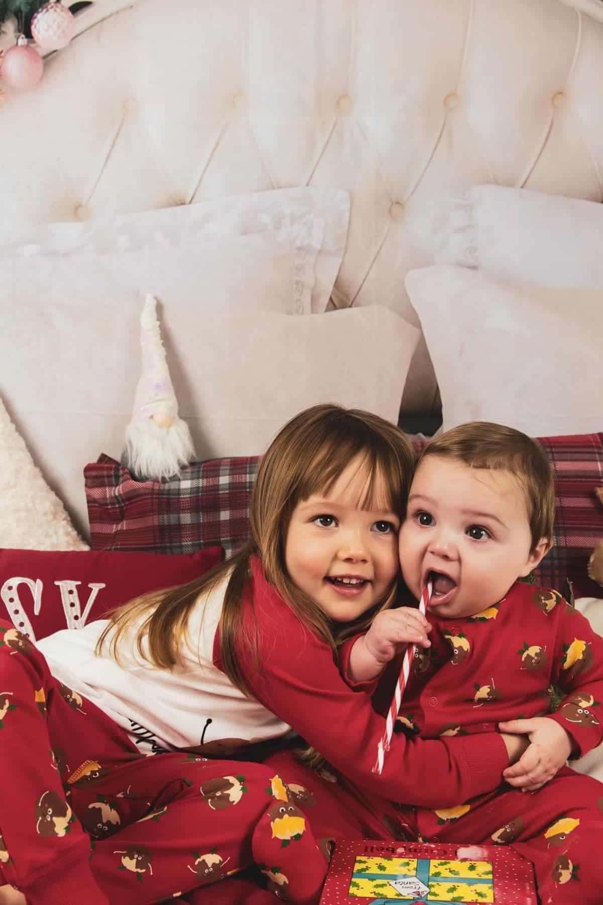 Two young children in matching red pajamas sit on a bed; the older child hugs the younger, who is holding a candy cane. Pillows and festive decor are visible in the background.