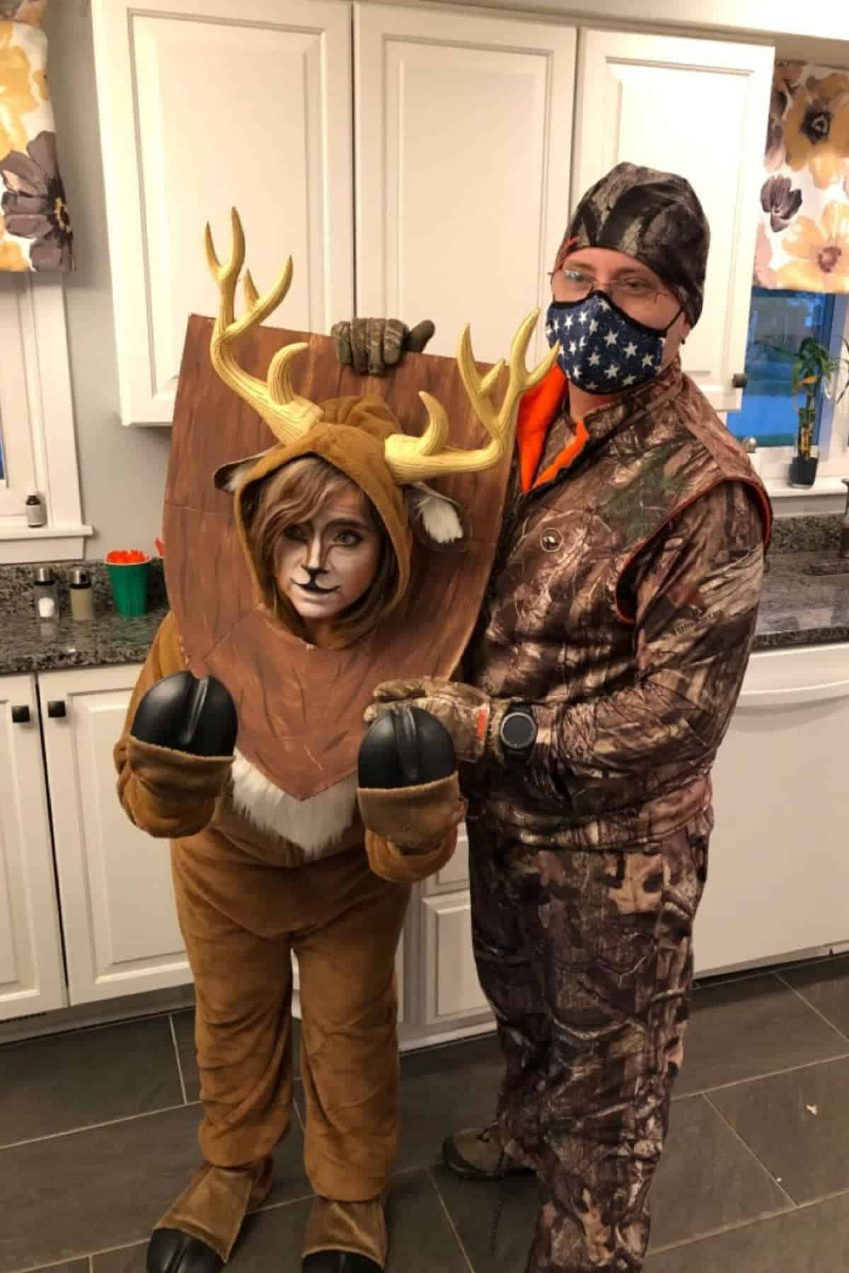 Two people in a kitchen; one is dressed as a deer with antlers and hooves, and the other is in camouflage with a star-spangled face mask.
