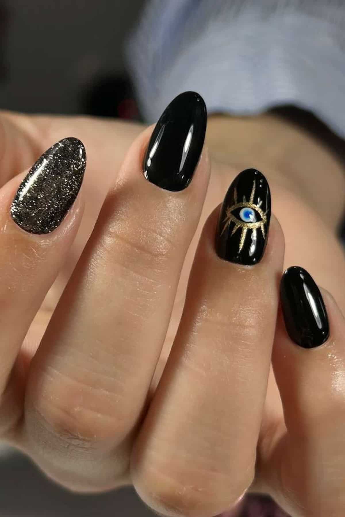 A hand with black and glitter nail polish; ring fingernail features a gold eye design with a blue center.