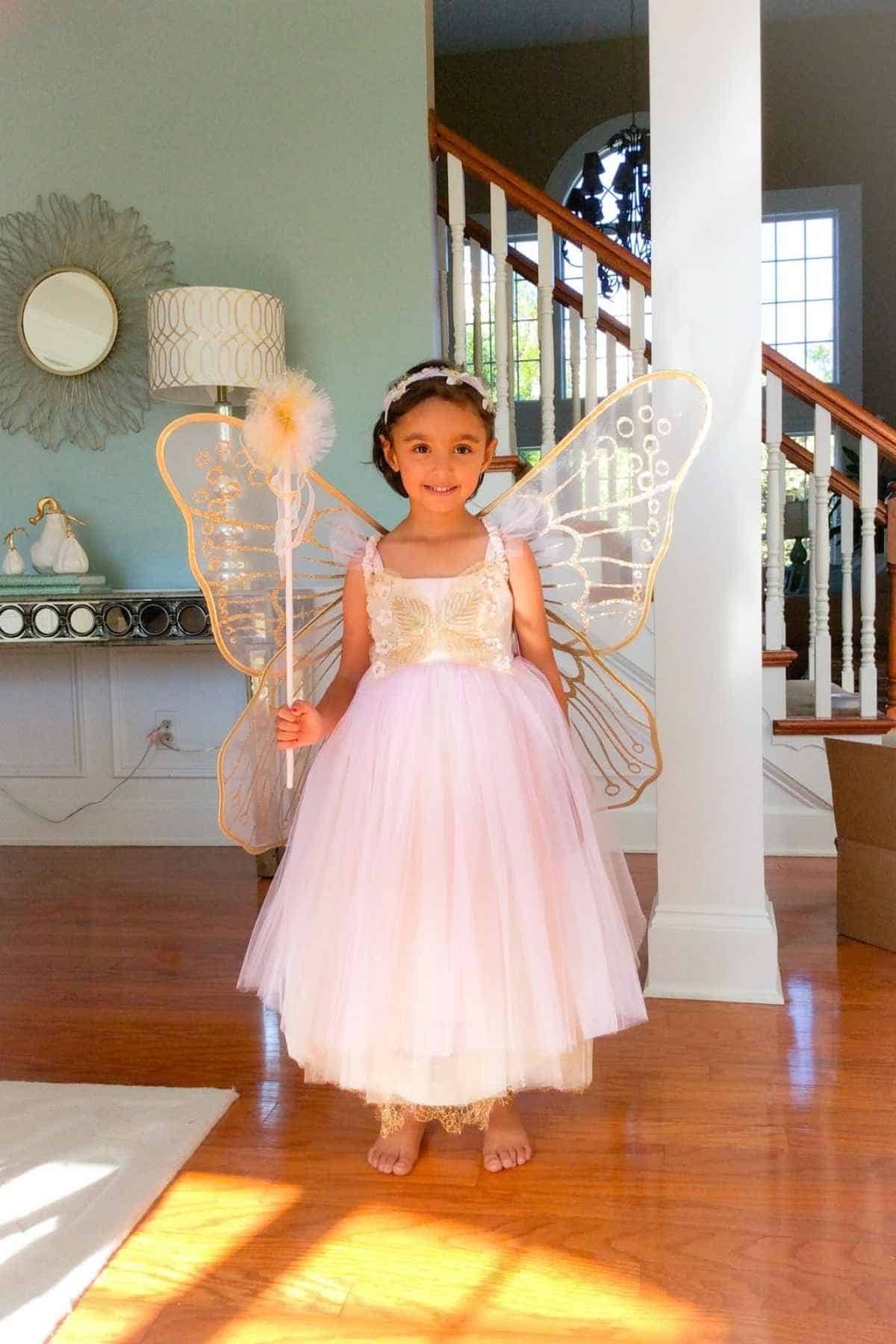 A young child in a fairy costume with wings and a wand stands indoors on a wooden floor, smiling at the camera.