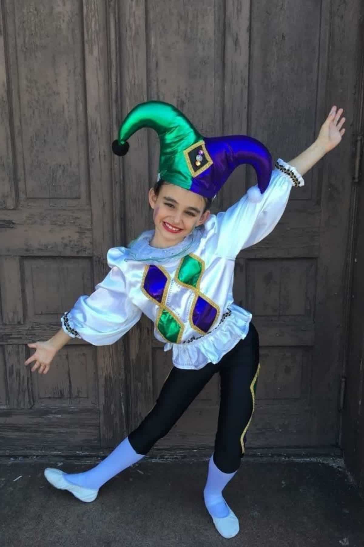 Child in a jester costume with a green and purple hat, posing with arms outstretched in front of a wooden door.