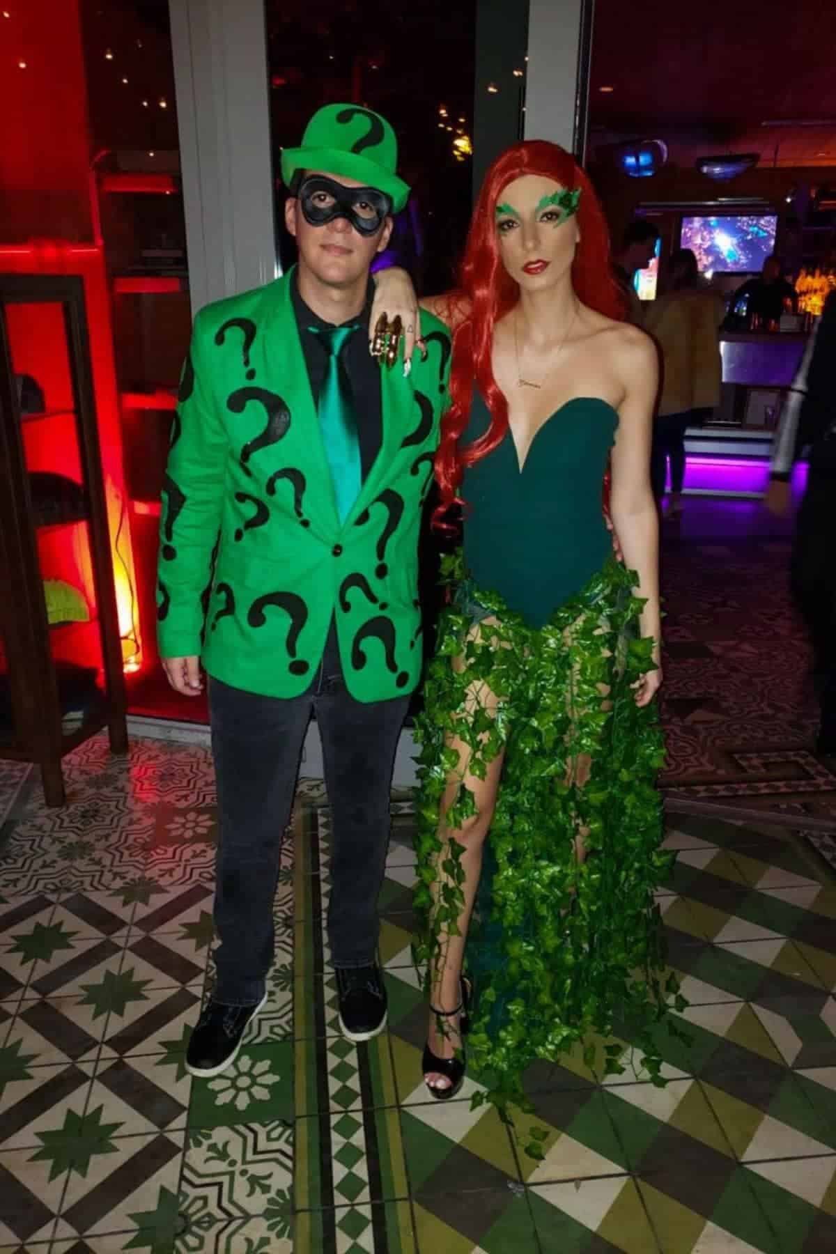 Two people in costumes: one dressed as the Riddler with a green suit and question marks, the other as Poison Ivy with a leafy green outfit and long red hair, standing indoors.