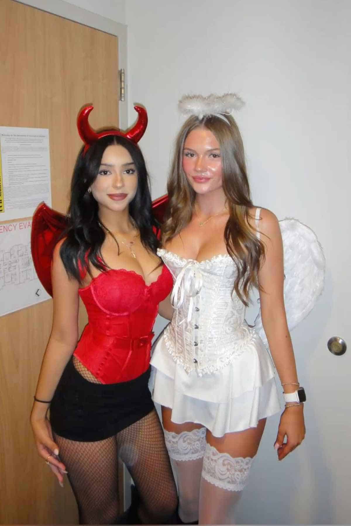 Two women pose indoors in costume; one wears a red devil outfit with horns and wings, while the other wears a white angel outfit with a halo and wings.