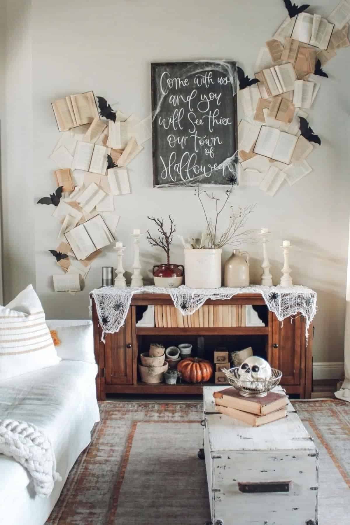 A living room decorated for Halloween with open books, black paper bats on the wall, pumpkins, a skull, branches, candles, and a sign reading “Come with us and we’ll see this our town of Halloween.”.