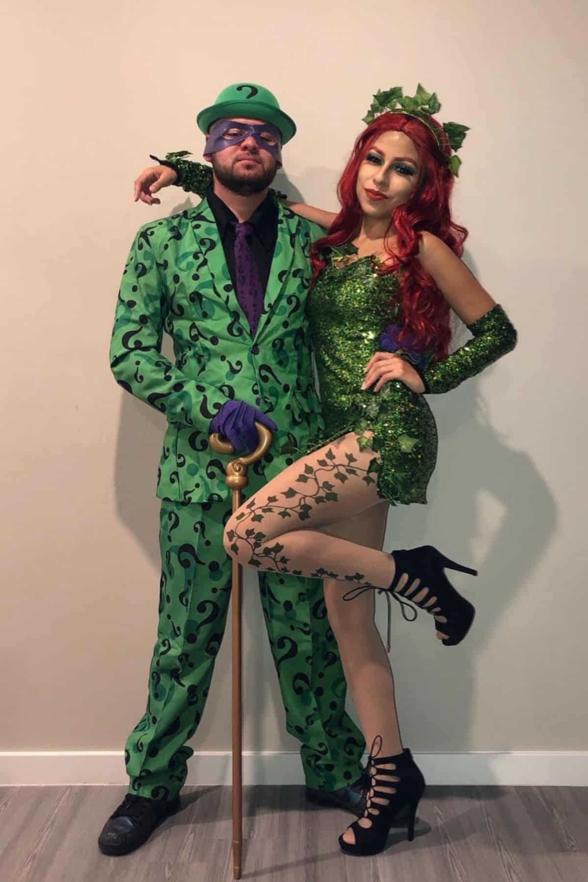 Two people in costumes: one dressed as the Riddler in a green suit with question marks, and one as Poison Ivy in a green dress with red hair and vine-patterned tights, posing together.