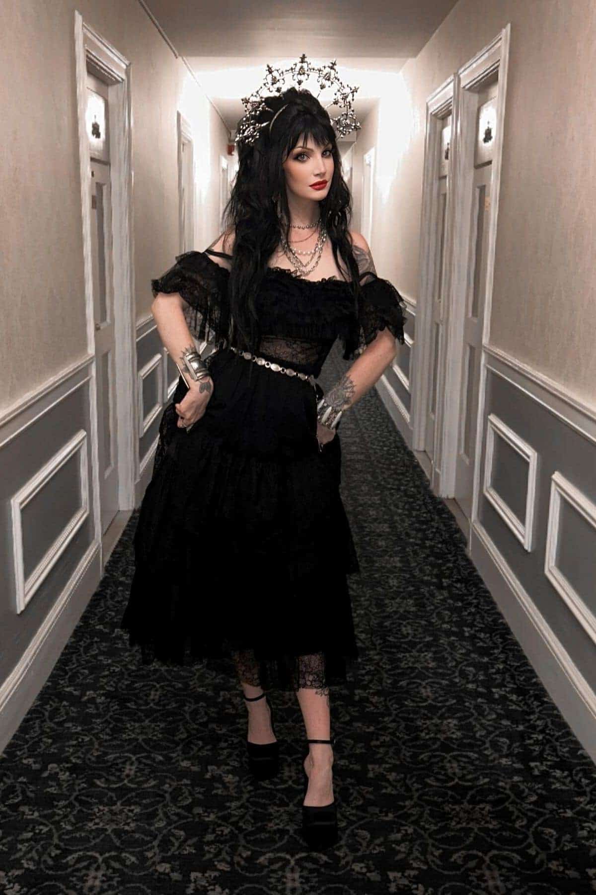 Woman in a black dress and crown stands in a carpeted hallway with white-paneled doors and beige walls, looking to the side.