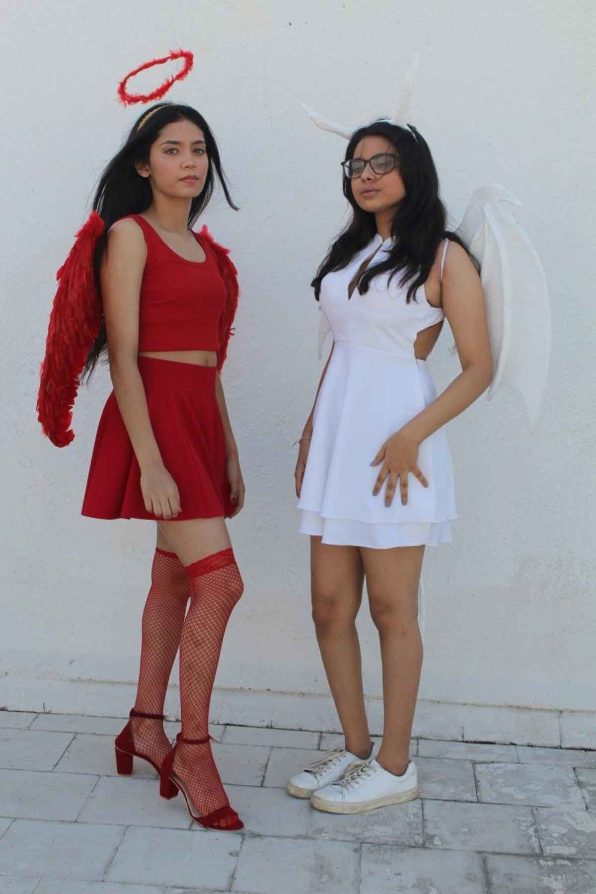 Two young women stand side by side; one wears a red angel costume with wings and halo, the other wears a white angel costume with wings, a headband, and glasses, against a plain wall.