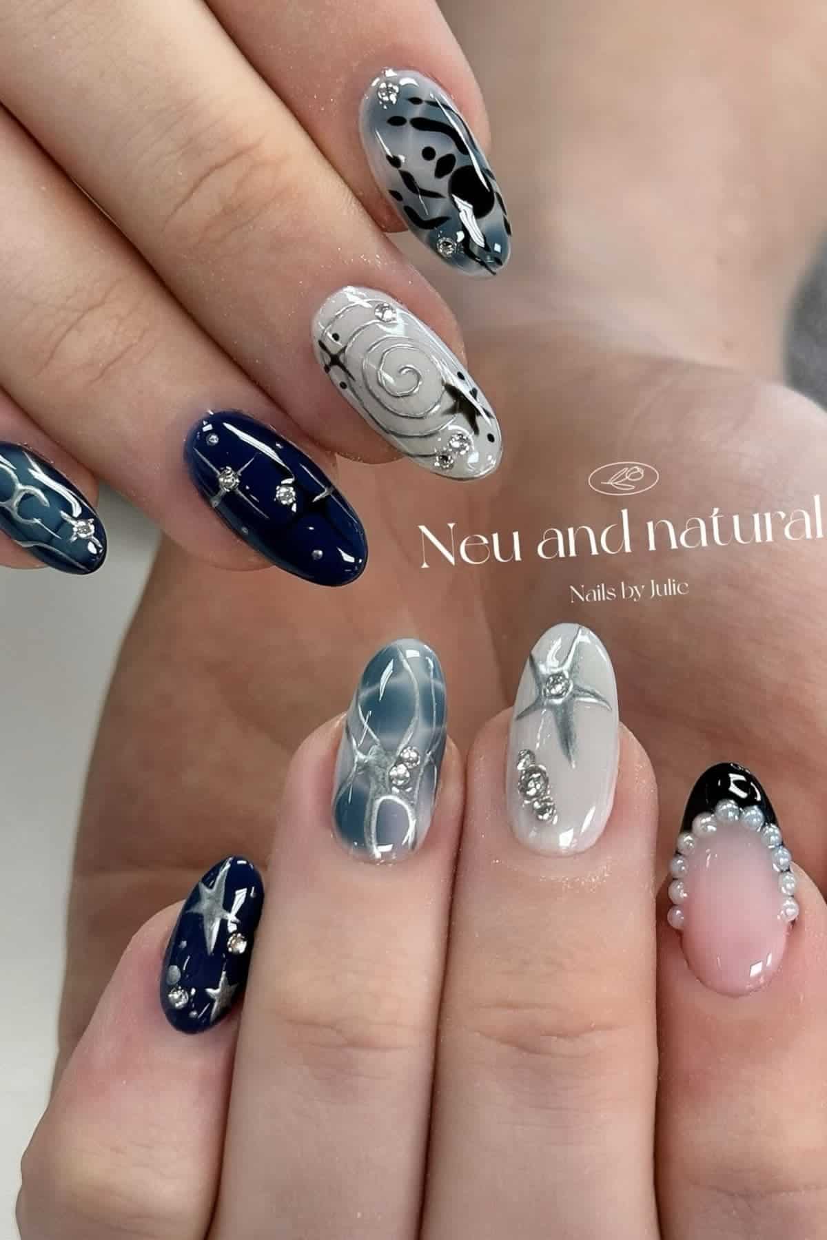 Close-up of hands with long, almond-shaped nails featuring various blue, white, and black abstract designs, rhinestones, and metallic accents.