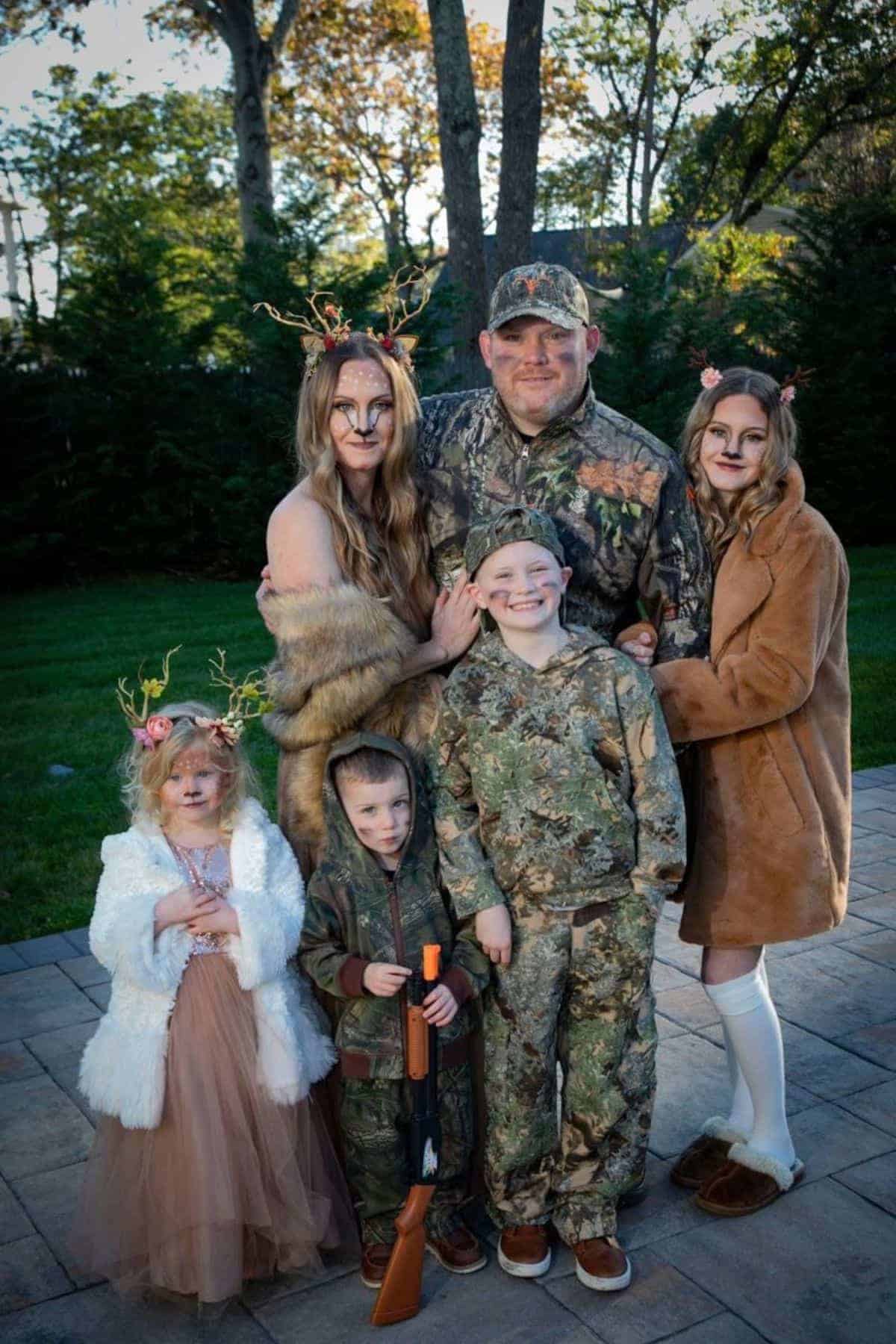 A family of six poses outdoors in coordinated woodland and deer costumes, with camo outfits and antler headbands, on a stone patio with trees in the background.