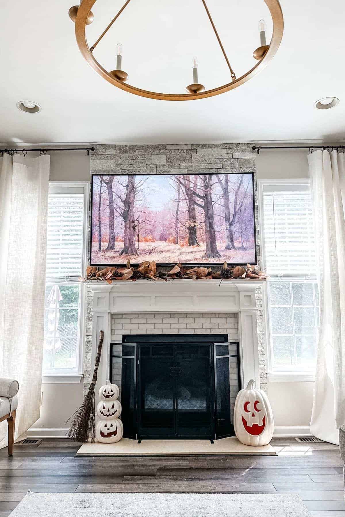 White fireplace with black screen, Halloween pumpkin decorations, broomstick, and a TV above displaying a forest scene. Light curtains frame two windows on either side.