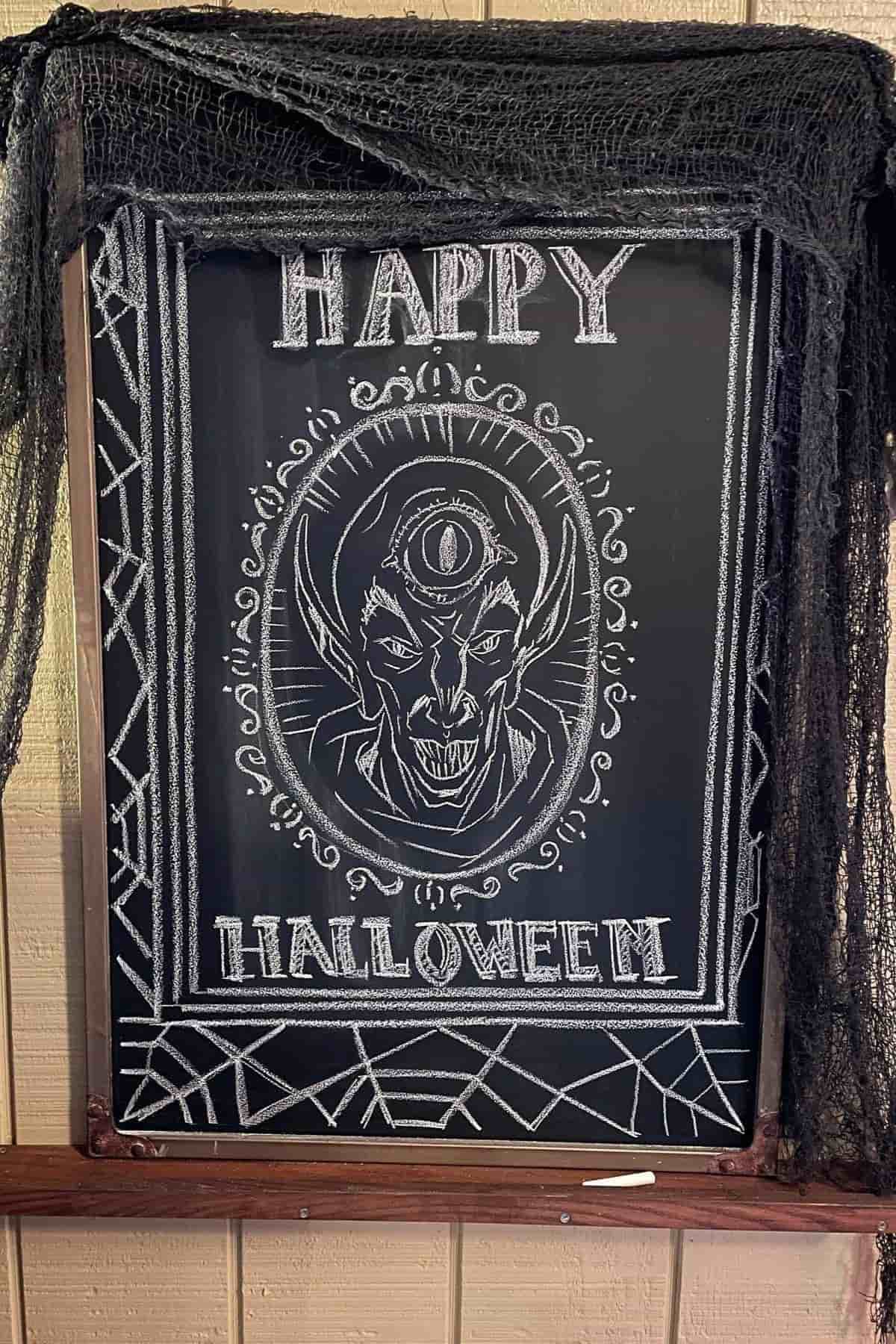 Chalkboard with “Happy Halloween” and a drawing of a vampire with a third eye, framed by geometric and decorative designs, draped with black netting.