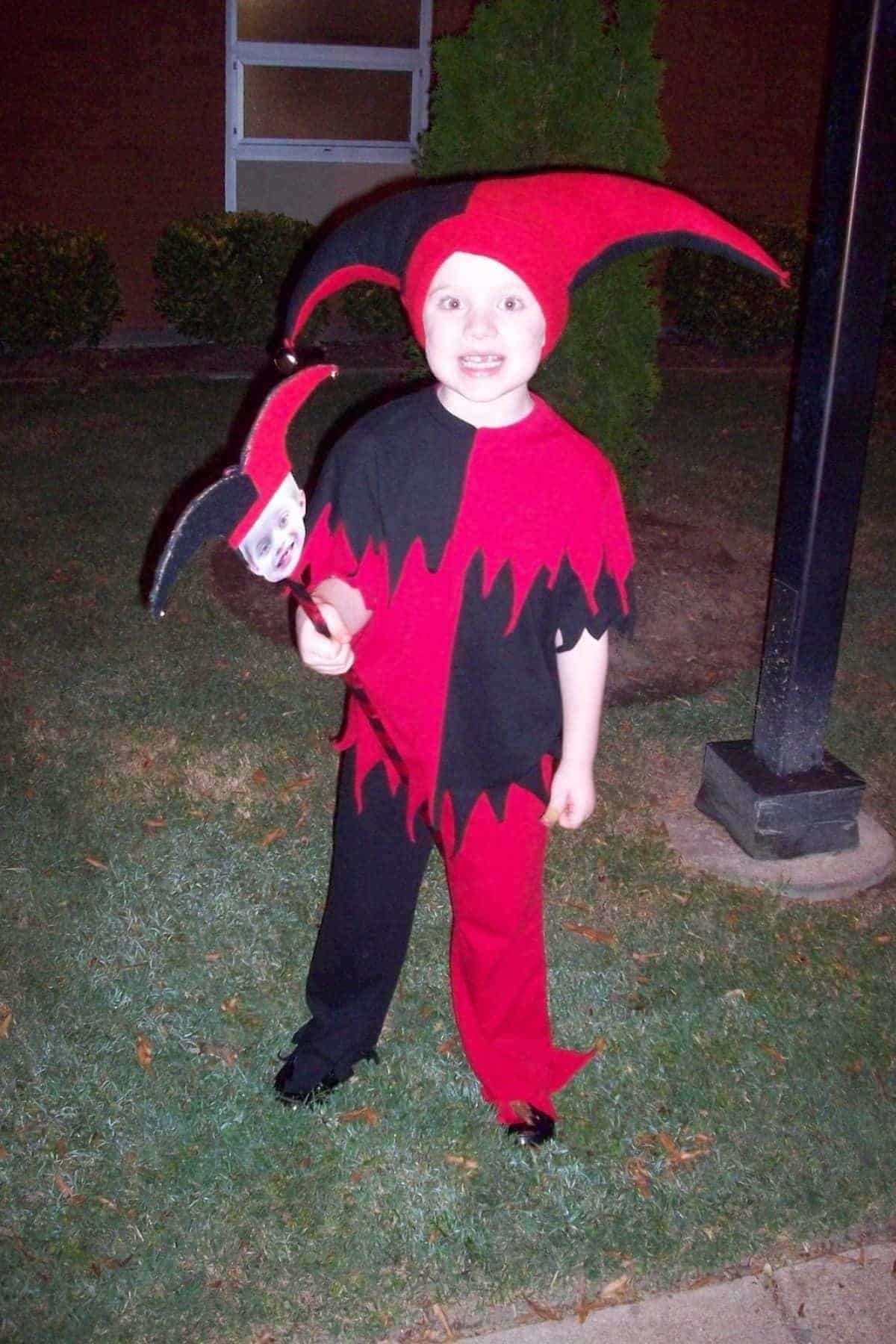 Child in a red and black jester costume stands on grass holding a matching jester stick with a face.