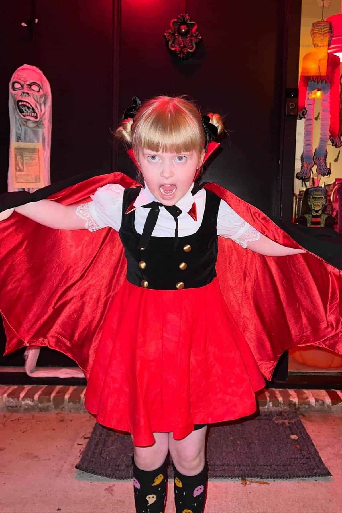Young child in a red and black vampire costume stands with arms outstretched, showing a cape, in front of a decorated door with Halloween decorations.