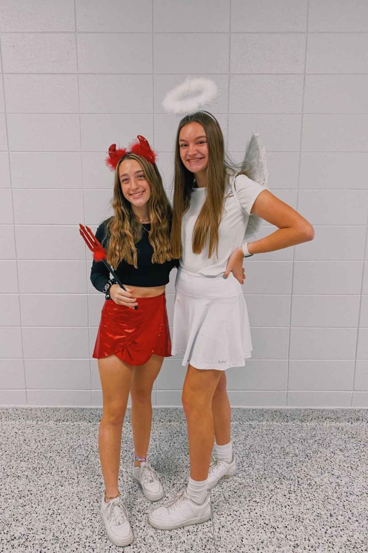 Two girls stand side by side; one is dressed as a devil in red, holding a trident, and the other is dressed as an angel with a halo and wings. Both are smiling in front of a tiled wall.