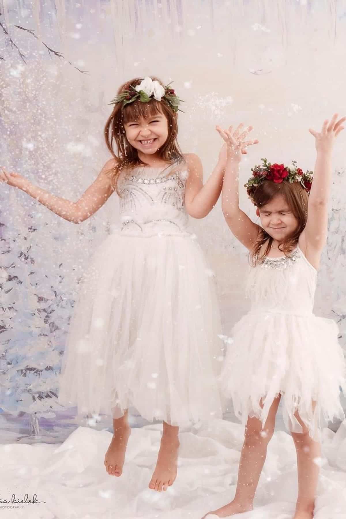 Two young girls in white dresses and floral crowns smile and play, tossing artificial snow against a winter-themed backdrop.