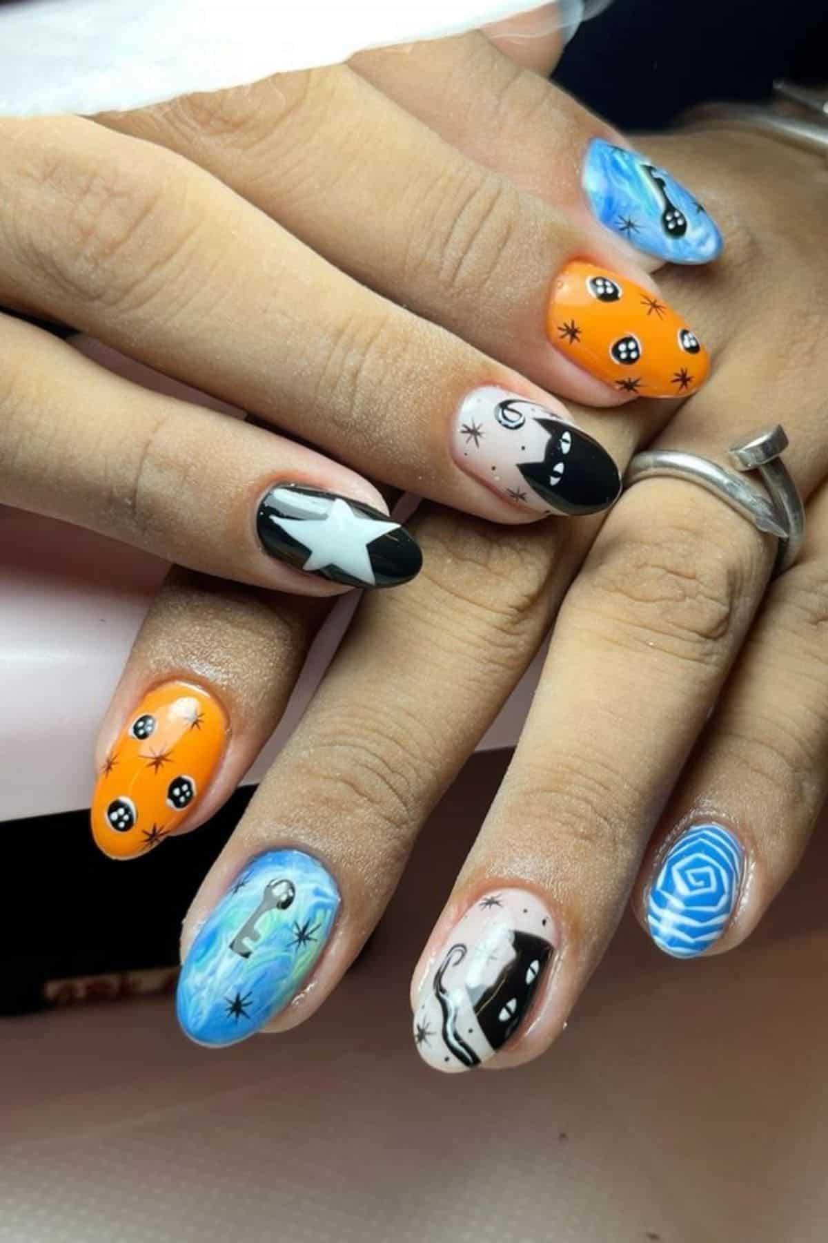 Hands with long nails featuring colorful designs, including stars, swirls, cat faces, orange and blue backgrounds, and abstract black-and-white shapes. The person is wearing two rings.