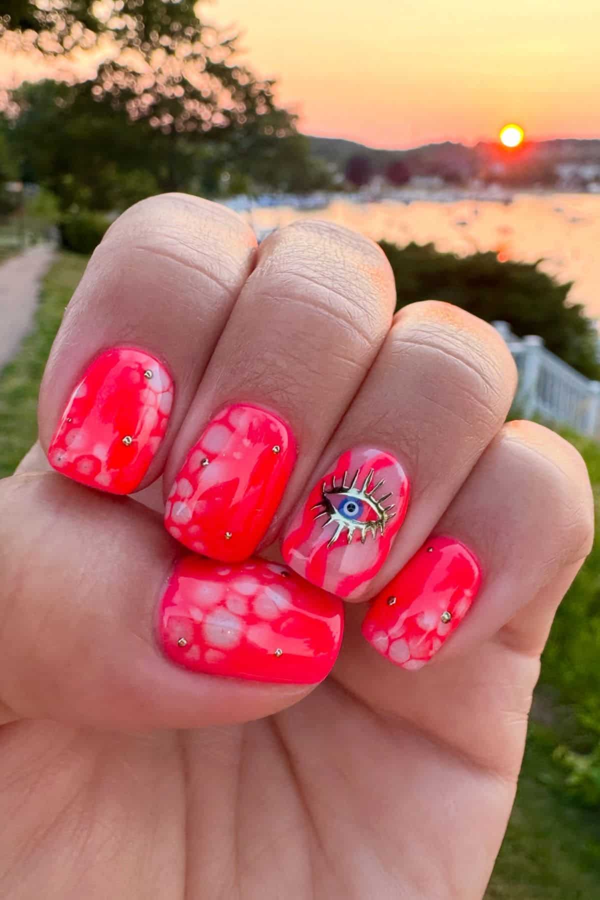 A hand with bright pink and white marbled nail art, featuring gold studs and an eye design on the ring finger, held outdoors at sunset near water.