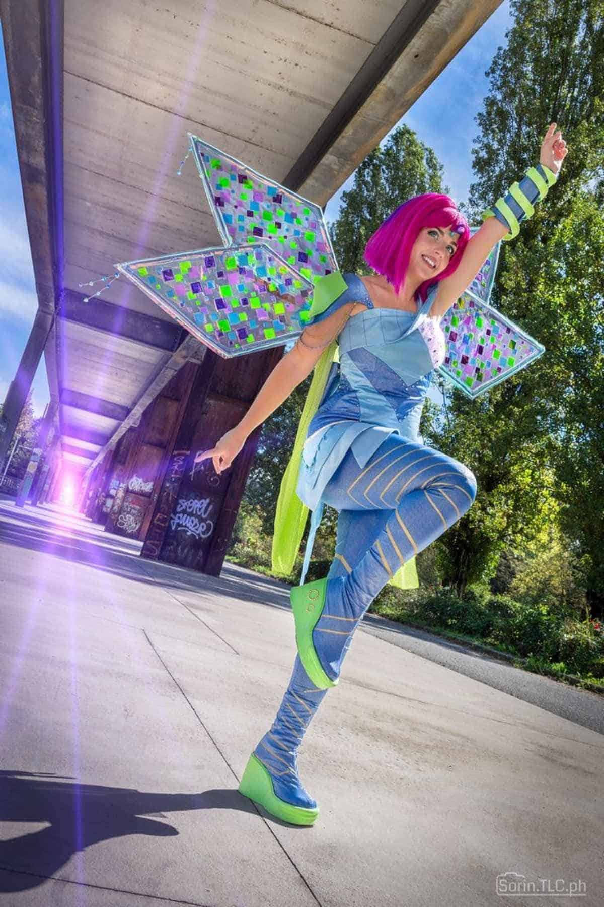 Person in colorful fairy costume with geometric wings, bright pink wig, and green platform shoes poses energetically outdoors under a covered walkway with sunlight in the background.