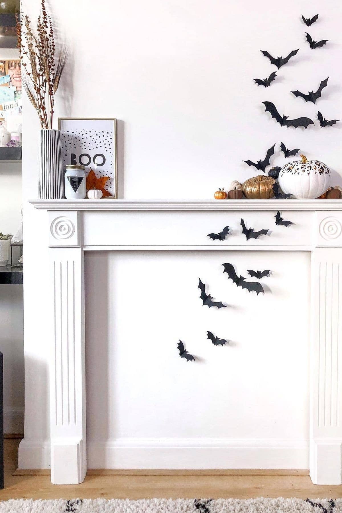 White mantel decorated with black paper bats, pumpkins, a vase with branches, and a framed “Boo” sign against a plain white wall.